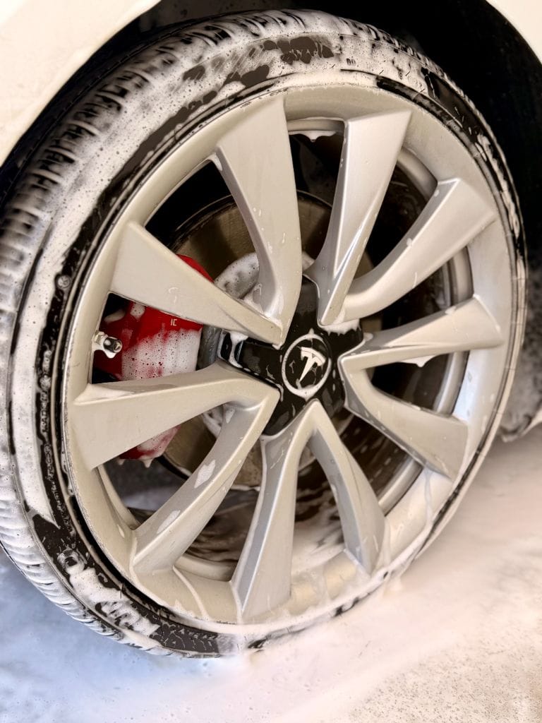 Close-up of a Tesla Model 3 wheel and red brake caliper covered in foam during a thorough wheel cleaning at Brightworks Fort Worth
